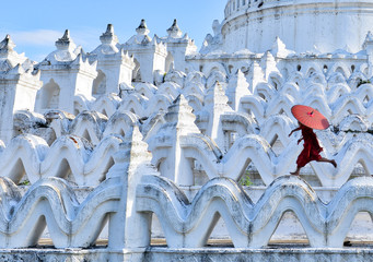 novice buddhist monks with red traditional robes holding red umbrellas walking in a white buddhist temple in myanmar