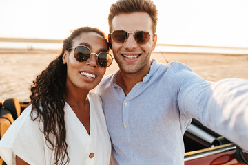 Photo of attractive multiethnic couple wearing sunglasses taking selfie photo together while standing by car on beach