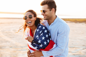 Photo of happy multiethnic couple wrapped in american flag smiling and hugging together by seaside