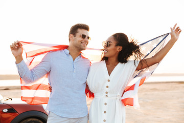 Photo of joyous multiethnic couple smiling and holding american flag while standing by car outdoors