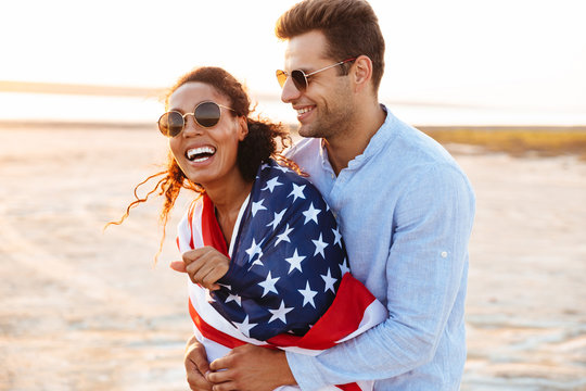 Photo of happy multiethnic couple wrapped in american flag smiling and hugging together by seaside - Powered by Adobe