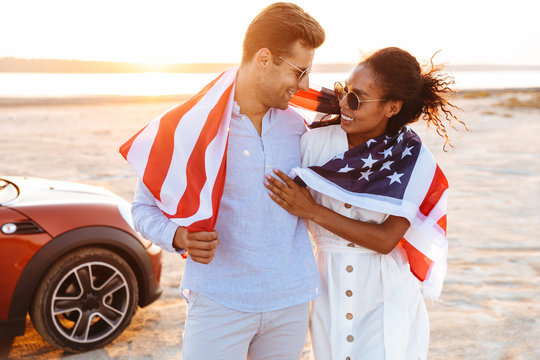 Photo of stylish multiethnic couple smiling and holding american flag while standing by car outdoors