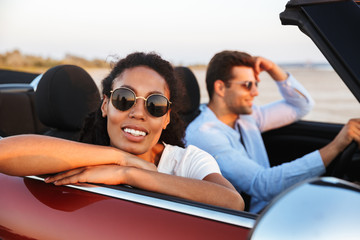 Image of beautiful multiethnic couple smiling while riding in convertible car by seaside at sunset