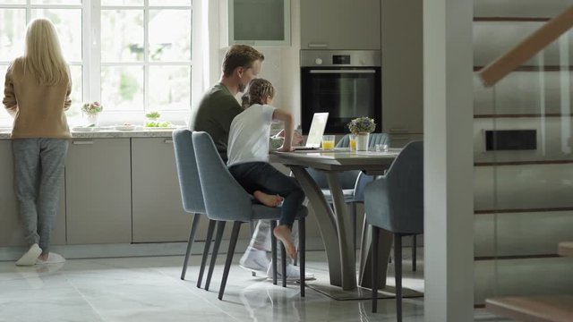 Tracking left shot of middle aged man browsing Internet on laptop sitting at table in domestic kitchen. Wife and little daughter joining him, family looking at laptop screen and doing online shopping