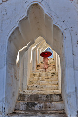 novice buddhist monks with red traditional robes holding red umbrellas walking in a white buddhist temple in myanmar