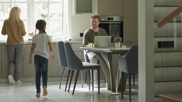 Tracking shot of man working on laptop sitting at table in domestic kitchen. Little daughter running downstairs to him, caring mother giving her glass of orange juice. Family gathering at table