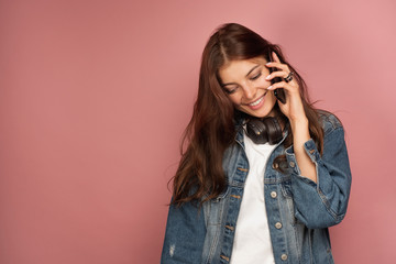 A girl in a jeans with headphones on her neck speaks on the phone on a pink background.