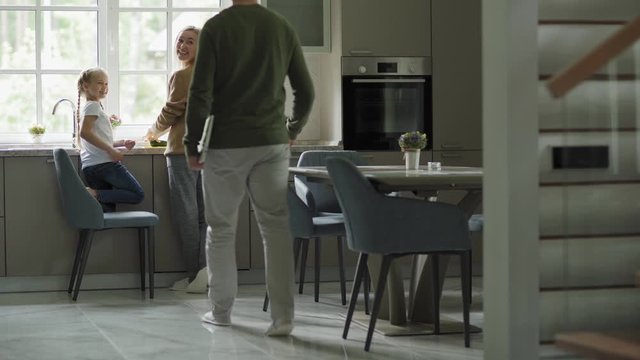 Mother and little daughter cooking together at kitchen counter. Father with laptop walking downstairs and working on computer at table