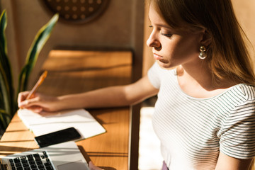 Portrait of charming young woman using laptop computer and writing notes in diary book in cafe