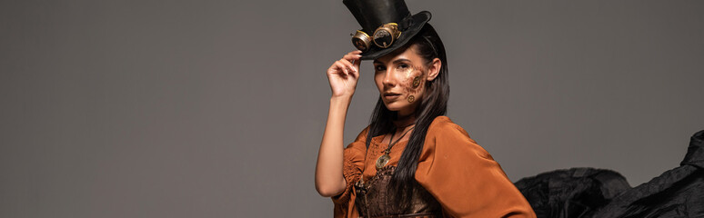 panoramic shot of attractive steampunk woman in top hat with goggles looking at camera isolated on...