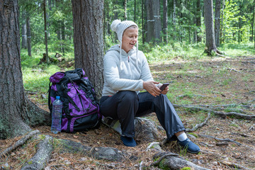 Obraz premium A pretty girl in light clothes, a European-looking ecotourist, sits near a tree in the forest and looks at the phone.