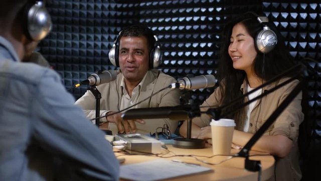 Tracking shot of unrecognizable radio host in headphones chatting with male Arab guest during recording of talk show in studio. Young Asian woman listening to them talk and smiling