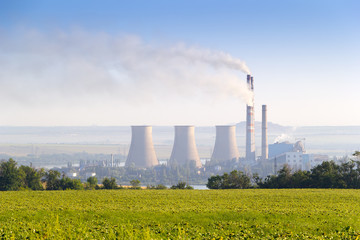 Environmental pollution concept. Pipes and towers of the thermoelectric plant. Green sunflowers field and tree planting in foreground.