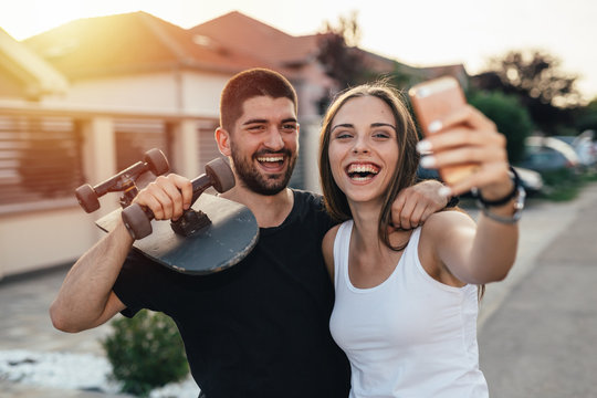 young modern couple walking embraced holding skateboard and taking picture with smartphone