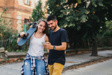 young happy couple holding arms, carrying a skateboard, using smartphone walking in the park