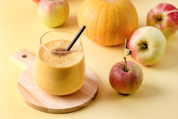 Glass of Fresh Tasty Pumpkin and Apples Juce or Smoothie with Cinnamon with Black Strew on Wooden Cutting Board Ripe Apples and Pumpkin on Yellow Background Horizontal