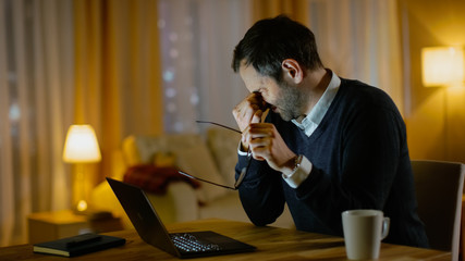Tired Middle Aged Man Works on a Laptop. He's Eyes Fatigued, He Temporarily Takes off His Glasses and Rubs His Eyes. His Apartment Looks cosy in Yellow Tones.