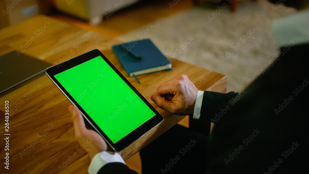 Poster at home man sits at his desk and holds tablet computer with green screen on it. his apartment is don