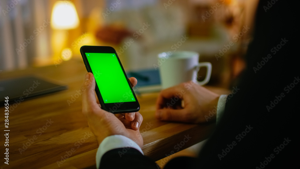Poster at home man sits at his desk and uses smartphone with green screen on it. he drinks from a cup. his 