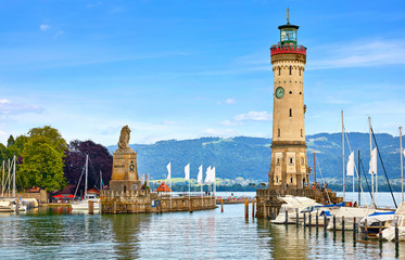 Lindau, Germany. Old lighthouse with clock in bay. Antique bavarian town at Lake Constance (Bodensee). Monument with statue of lion at entrance to port, yachts by piers. Summer landscape blue sky. - obrazy, fototapety, plakaty