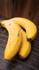 Three ripe Cavendish bananas with small bruises showing ripeness positioned on a wooden brown table, healthy fruit popular for its fiber, potassium and magnesium content; vertical, narrow perspective