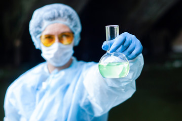 woman in medical or laboratory suit shows flask with greenish liquid on a dark background