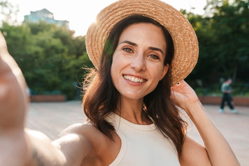 Image of nice middle-aged woman taking selfie photo and looking at camera while walking in summer park