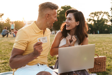 Portrait of joyful middle-aged couple holding credit card while using laptop computer during picnic in summer park