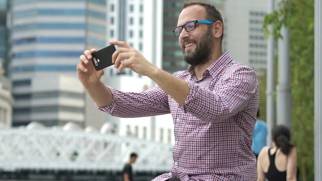 Young, happy man taking selfie with cellphone sitting in city park