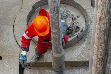 A working driller in a red uniform, in a helmet and goggles, installs drill pipes after lifting...