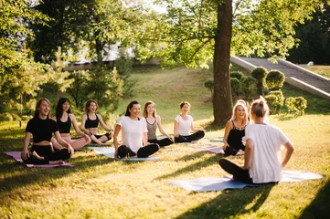 Foto op Aluminium Groep glimlachende vrouwen bereiden zich voor op meditatie in het park op zonnige zomerochtend onder begeleiding van een trainer. Groep meisjes zitten in lotushouding op yogamatten ontspannen praten voor yogatraining © dikushin