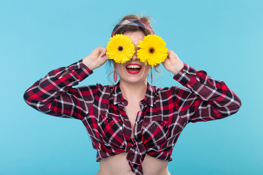 Close-up positive smiling young woman in a red checkered shirt holding yellow flowers on her eyes posing against a blue background. Concept incognito and fun.