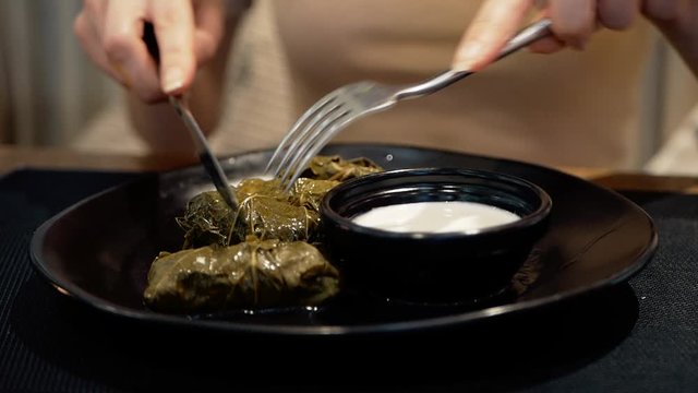 National traditional cuisine food in the restaurant. Woman is eating dolma with sour cream served in clay plate, dish closeup. Girl is eating with fork and knife
