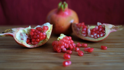 One whole and one open pomegranate or the fruit of Punica granatum revealing clusters of red, juicy seeds, white fibrous arils and thick, waxy peel resting on a wooden board with a burgundy background