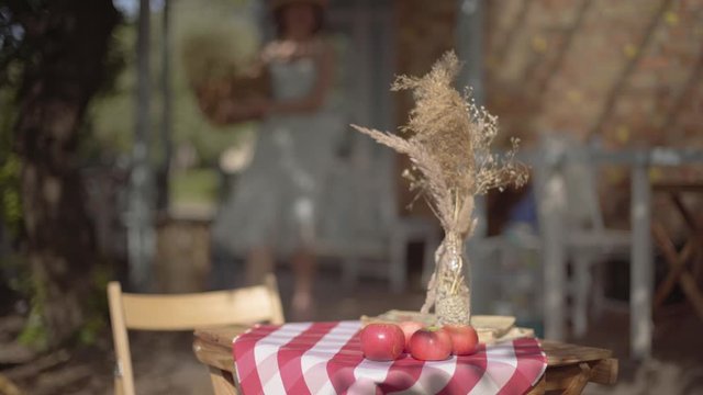 Blurred figure of a young rural girl walking with the wicker basket in the background. Small table with apples and vase with decorative grass in it in the foreground. Rural lifestyle