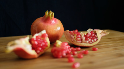 One whole and one open pomegranate or the fruit of Punica granatum revealing clusters of red, juicy seeds, white fibrous arils and thick, waxy peel resting on a wooden board with a dark background