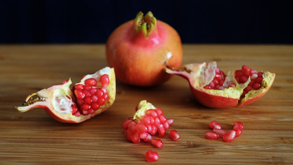 One whole and one open pomegranate or the fruit of Punica granatum revealing clusters of red, juicy seeds, white fibrous arils and thick, waxy peel resting on a wooden board with a dark background