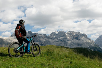 Fototapeta na wymiar man with electric bike, e-bike, ebike, looking mountains of Brenta Peak, Grostè Pass, meadow, Dolomites, Madonna di Campiglio, summer, sport, adventure, travel, Alps, Trentino Alto Adige, Italy