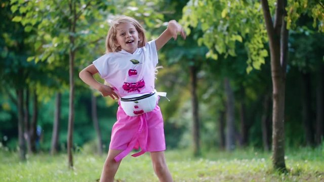 Positive little girl dancing happily in the park. Smiling child in summer suit having fun outdoors. Slow motion.