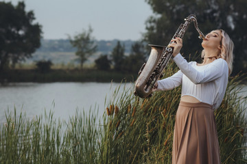 Portrait of young musician on nature background, beautiful woman playing saxophone on bank river in...