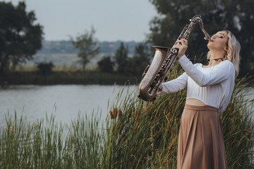 Portrait of young musician on nature background, beautiful woman playing saxophone on bank river in...