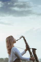 profile of a young beautiful girl playing the saxophone against a cloudy sky, a woman relaxes on nature, music and hobby concept