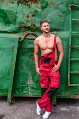 An einem alten grünen Container lehnender muskulöser junger Mann in einer roten Latzhose.