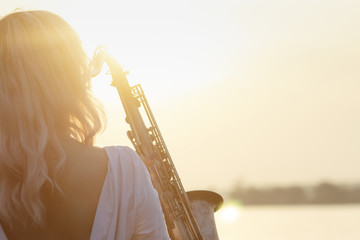 silhouette of a young beautiful girl playing the saxophone at sunrise by the river, woman on the...
