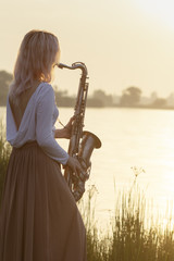 silhouette of a young beautiful girl playing the saxophone at sunrise by the river, a woman in a...