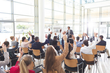Business people listening to businessman and raising hands to ask him questions at conference.