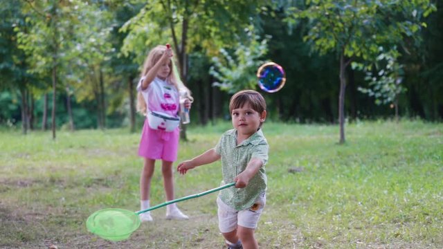 Little children playing with soap bubbles in nature. Pretty girl blowing colorful bubbles and small cute boy trying to catch them in the park.