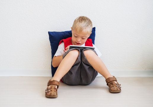 A portrait of down-syndrome school boy sitting on the floor, using tablet.