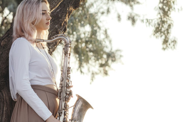 portrait of a young beautiful girl holding a saxophone leaning on a tree trunk, blonde woman...