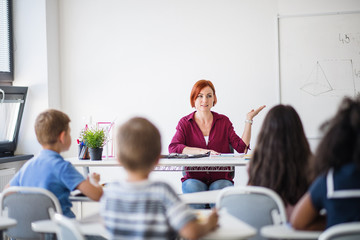 Naklejka na ściany i meble Rear view of school children sitting at the desk in classroom, listening to teacher.